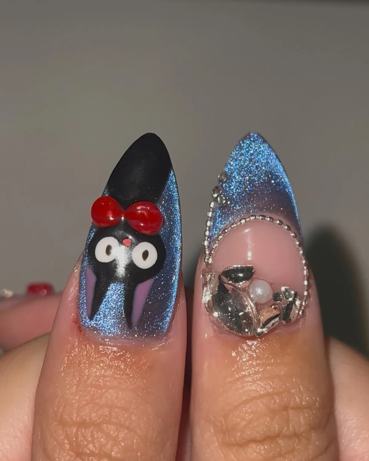 Jiji the black cat with a red bow on a blue cat-eye nail, next to a nail with crystals and a pearl